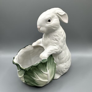 Puede incluir: Una figura de conejo de cerámica blanca de pie con sus patas delanteras apoyadas en el borde de un cuenco hueco con forma de repollo verde y blanco. El conejo tiene un acabado liso, nariz rosa y un ojo oscuro, mientras que el repollo tiene hojas texturizadas.