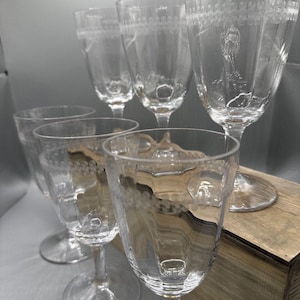 Op de afbeelding: Een set van vijf heldere glazen op een voet met een ronde basis. Elk glas is voorzien van een delicaat geëtst patroon van herhalende krullen en lijnen rond de bovenrand, met een gladde, transparante kelk en een stevige, heldere glazen steel.