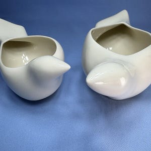 May include: Two off-white ceramic bird-shaped bowls with smooth, glossy finishes. Each vessel features a pointed beak-like protrusion at the front and a curved tail-like extension at the back, creating a hollow, open-top container design.