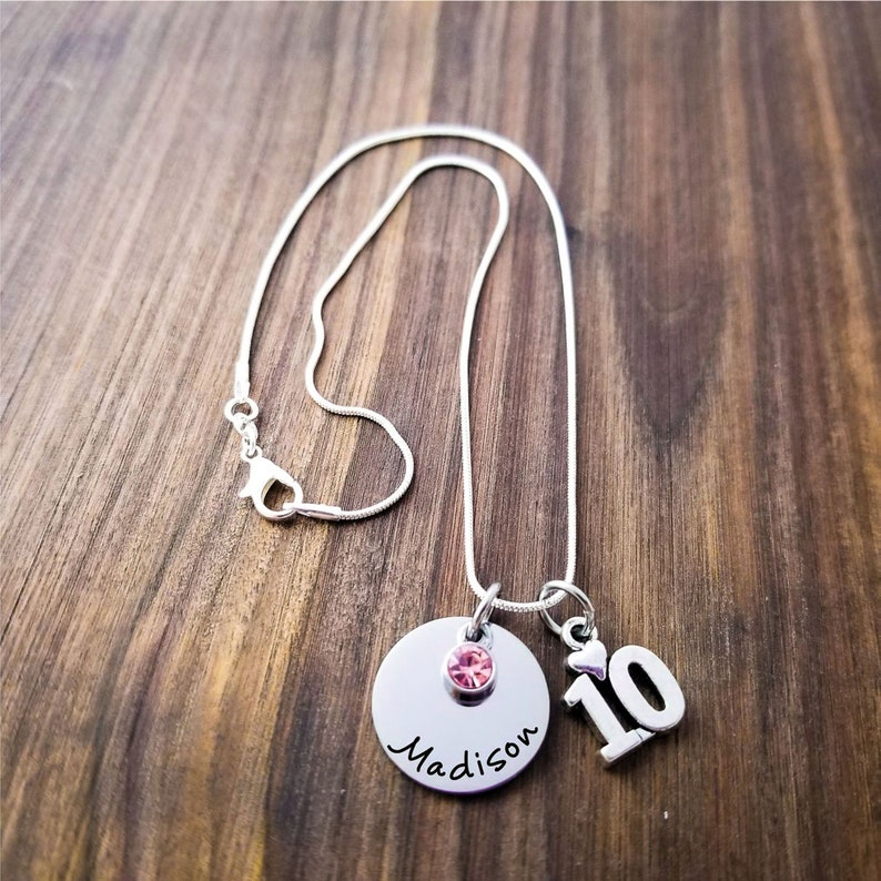 May include: A silver necklace with a round pendant, a pink gemstone, and the name "Madison" in cursive. A charm with the number "10" is also attached. The necklace is displayed on a wooden surface.