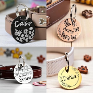 May include: Four round pet ID tags with different designs. One black tag has the name "Daisy" with a butterfly and floral design. A rose gold tag has the name "Annie" with butterflies. A silver tag has "Sunny" with a beach scene. A gold tag has "Dahlia" with floral accents.