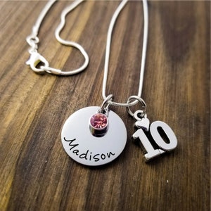 May include: A silver necklace with a pendant featuring the name "Madison" in cursive, a pink gemstone, and a charm with the number "10". The necklace is displayed on a wooden surface.