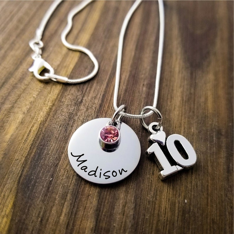 May include: A silver necklace with a pendant featuring the name "Madison" in cursive, a pink gemstone, and a charm with the number "10". The necklace is displayed on a wooden surface.