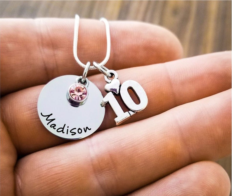 May include: Silver-toned necklace with a round pendant engraved with the name "Madison" in cursive. A pink gemstone and a charm with the number "10" are attached. The necklace is displayed against a blurred background.