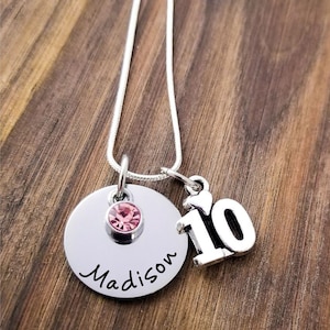 May include: A silver necklace with a round pendant featuring the name "Madison" in cursive and a pink gemstone. A charm with the number "10" hangs next to the pendant. The necklace is on a wooden surface.