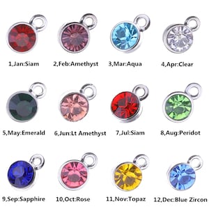 May include: A collection of twelve round gemstone charms in various colors, each with a silver-colored loop. The charms are labeled with months and gemstone names, including Siam, Amethyst, Aqua, Clear, Emerald, Lt Amethyst, Peridot, Sapphire, Rose, Topaz, and Blue Zircon.