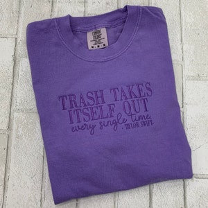 May include: A purple Comfort Colors t-shirt with the embroidered text "TRASH TAKES ITSELF OUT every single time - Taylor Swift." The shirt is folded and lies on a light-colored brick surface.