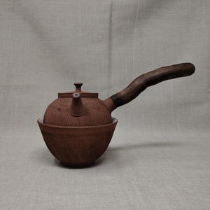 Könnte beinhalten: Eine braune Keramik-Teekanne mit Holzgriff. Die Teekanne hat einen runden Körper und einen Ausguss. Der Griff ist nach oben gebogen. Die Teekanne steht in einer passenden Schale. Der Hintergrund ist neutral.