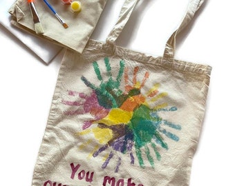 May include: A white canvas tote bag with a rainbow handprint design and the text "You make our heart shine". The bag comes with a set of paint and brushes.