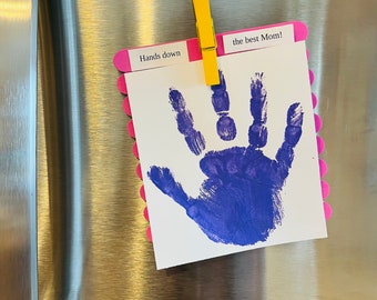 May include: A purple handprint on a white card with pink popsicle sticks around the edges. The card is attached to a refrigerator with a yellow clothespin. The card says "Hands down the best Mom!"