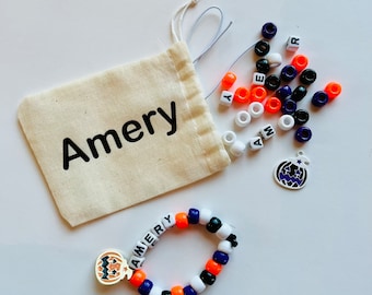 May include: A white drawstring bag with the name "Amery" printed on it. The bag contains a variety of colorful beads, including orange, white, black, and purple. There is also a small pumpkin charm with a black face and a star on top.