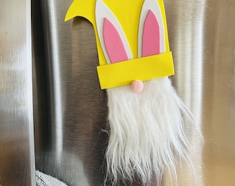 May include: A yellow felt gnome with pink bunny ears and a white fuzzy beard. The gnome is attached to a silver refrigerator.