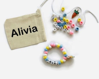 May include: A white canvas bag with the name "Alivia" printed on it. The bag contains a set of colorful alphabet beads, a string, and a carrot charm. A completed bracelet with the name "AMERY" is also shown.