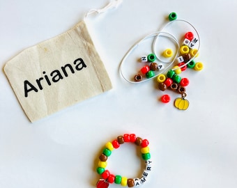 May include: A white drawstring bag with the name "Ariana" printed on it. Next to the bag are red, green, yellow, and brown beads with white letter beads spelling "Merry" and a small pumpkin charm.
