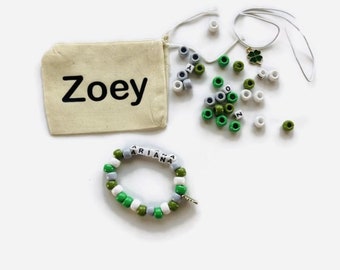 May include: A white drawstring bag with the name "Zoey" printed on it. The bag contains green, white, and gray beads with letters and a four-leaf clover charm. A bracelet made with the beads spells out the name "Ariana".
