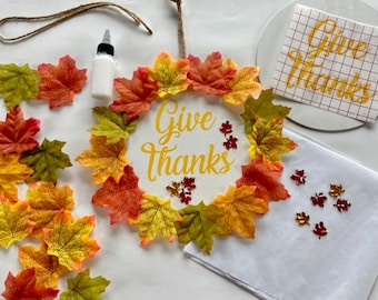 May include: A craft project to make a Thanksgiving wreath. The wreath is made of a white circle with the words "Give Thanks" in yellow lettering. The circle is surrounded by a ring of fall-colored leaves. There are also some small gold glitter leaves and a bottle of glue.