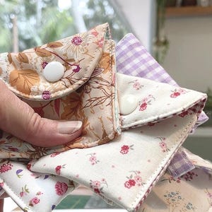 May include: A collection of small, handmade fabric pouches in assorted floral and patterned designs. The pouches are held together, displaying the different fabrics, including a floral print, a gingham pattern, and a cream fabric with small pink flowers. Each pouch has a white button closure.