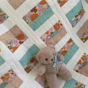 May include: A handmade quilt with a patchwork design featuring floral patterns in shades of pink, orange, and blue, interspersed with solid light blue and beige stripes. A small, plush teddy bear with a blue and white striped bow rests on the quilt.