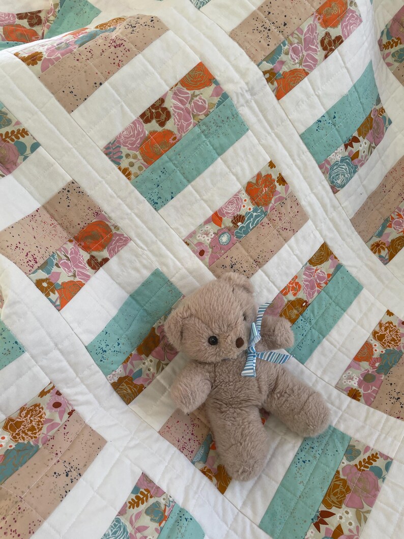 May include: A handmade quilt with a patchwork design featuring floral patterns in shades of pink, orange, and blue, interspersed with solid light blue and beige stripes. A small, plush teddy bear with a blue and white striped bow rests on the quilt.