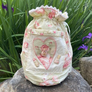 May include: A pale yellow fabric drawstring bag with a heart-shaped patch featuring a pink rose and a button. The bag has a floral print with pink roses and is decorated with pink buttons.