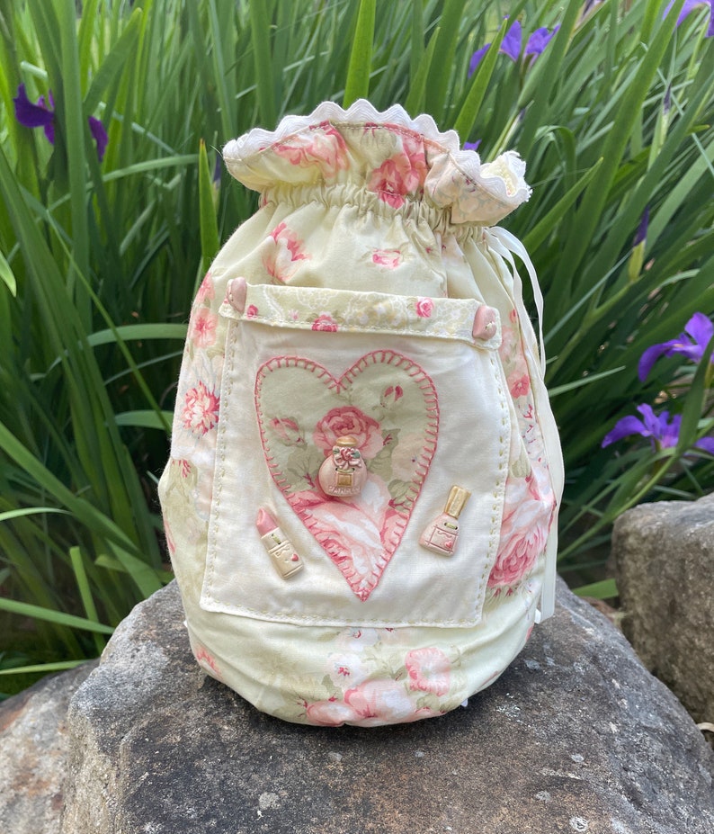 May include: A pale yellow fabric drawstring bag with a heart-shaped patch featuring a pink rose and a button. The bag has a floral print with pink roses and is decorated with pink buttons.