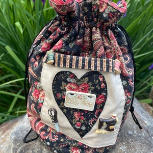 May include: A black and white fabric drawstring bag with a heart-shaped patch that says "Quilt Shop". The bag has a floral pattern and a red trim. There are sewing-themed embellishments on the bag, including a pair of scissors, a sewing machine, and spools of thread.