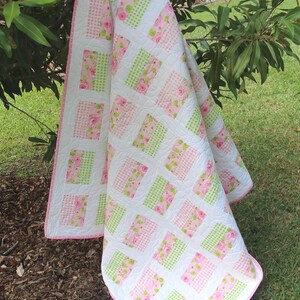 May include: A handmade quilt with a white background and a pattern of pink floral and green checkered squares. The quilt is draped over a tree branch on a grassy lawn. The quilt has a pink border.