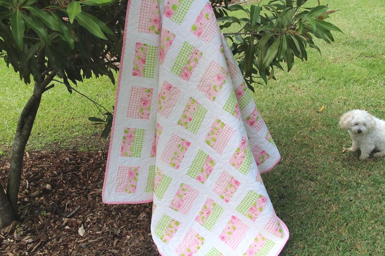 May include: A handmade quilt with a white background and a pattern of pink floral and green checkered squares. The quilt is draped over a tree branch on a grassy lawn. The quilt has a pink border.