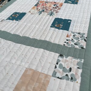 May include: A white quilted table runner with a patchwork design featuring various floral and geometric patterns in shades of green, beige, and pink.
