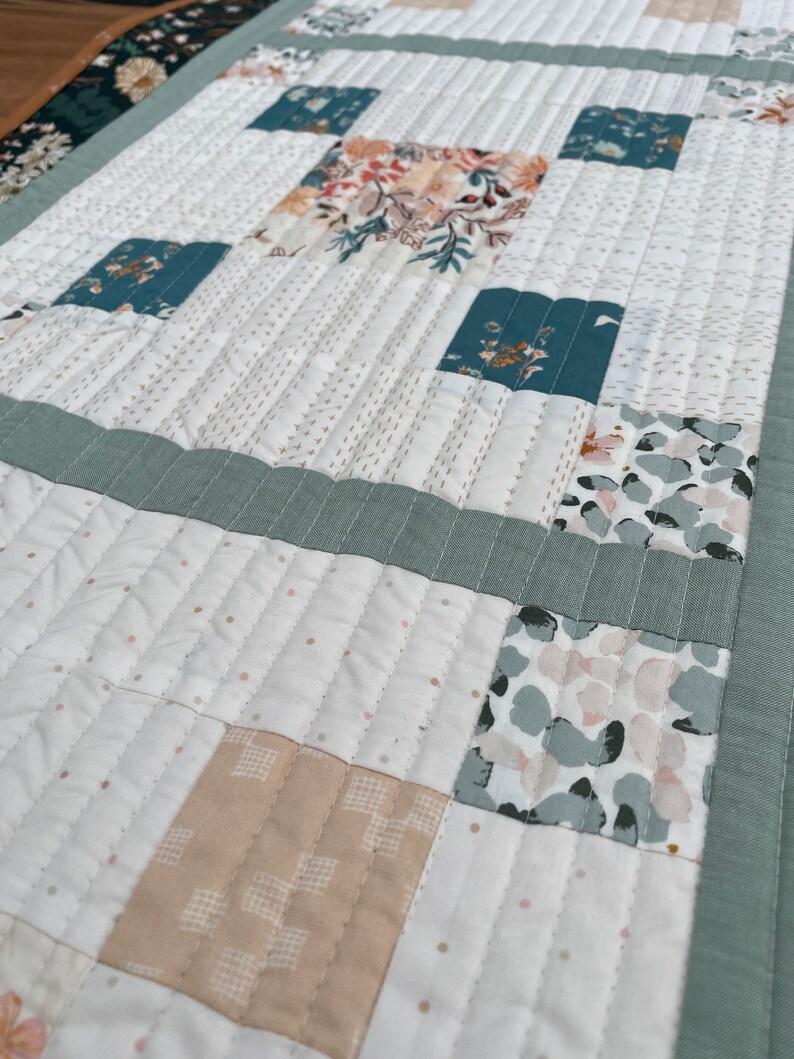 May include: A white quilted table runner with a patchwork design featuring various floral and geometric patterns in shades of green, beige, and pink.