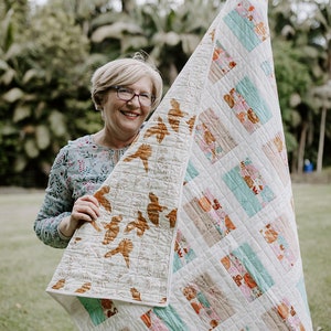 May include: A woman holds up a white quilt with a patchwork design featuring brown, pink, and teal colors. The quilt has a floral pattern and is made of different fabrics.