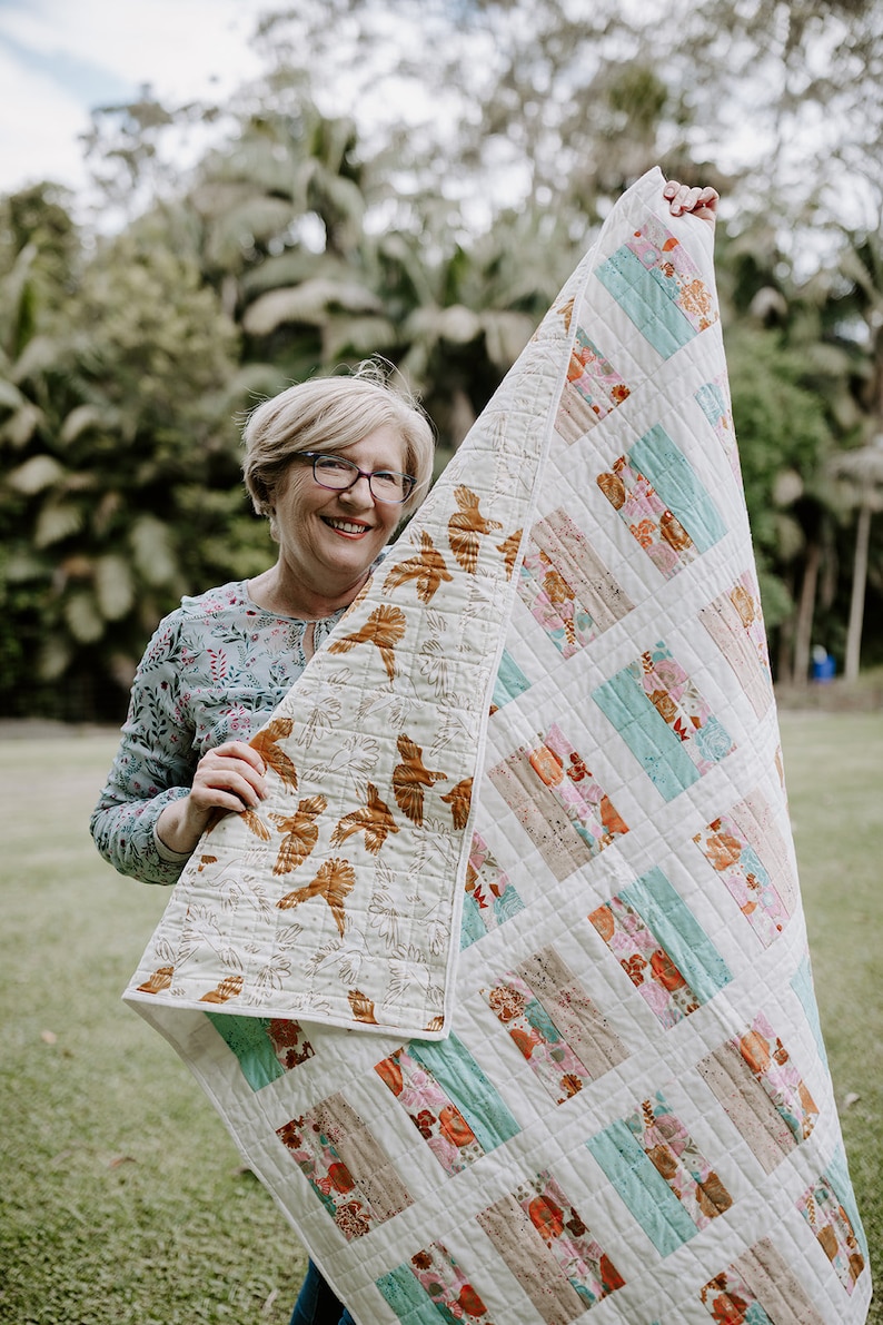 May include: A woman holds up a white quilt with a patchwork design featuring brown, pink, and teal colors. The quilt has a floral pattern and is made of different fabrics.