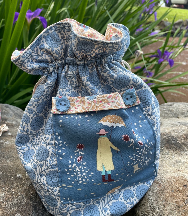 May include: A blue and white drawstring bag with a floral pattern. The bag has a pocket with a scene of a person walking a dog in the rain. The pocket has two blue buttons.