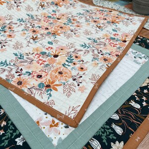 May include: Three quilted table runners with floral patterns. The top runner features a white background with orange, yellow, and green floral designs. The middle runner has a white background with pink and orange floral designs. The bottom runner has a black background with white and green floral designs. All runners have brown and green trim.