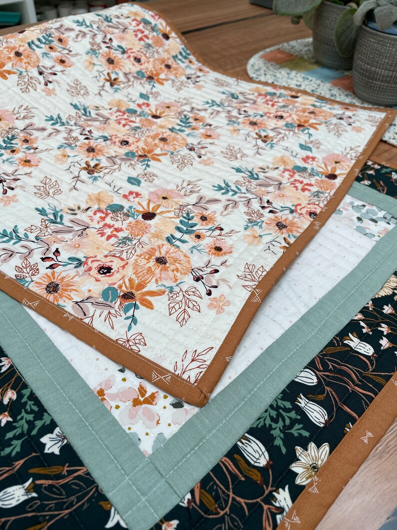 May include: Three quilted table runners with floral patterns. The top runner features a white background with orange, yellow, and green floral designs. The middle runner has a white background with pink and orange floral designs. The bottom runner has a black background with white and green floral designs. All runners have brown and green trim.