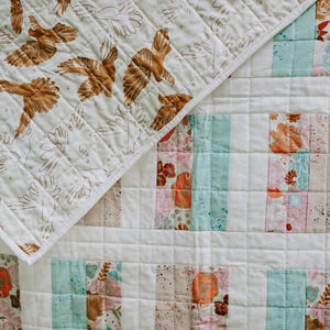 May include: A quilted blanket with a patchwork design. The top corner features a cream-colored fabric with brown bird and floral patterns. The main body has vertical stripes of floral, teal, and pink fabrics, creating a cozy, handmade aesthetic.