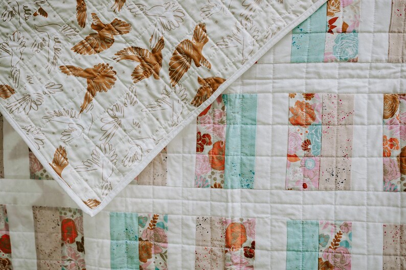 May include: A quilted blanket with a patchwork design. The top corner features a cream-colored fabric with brown bird and floral patterns. The main body has vertical stripes of floral, teal, and pink fabrics, creating a cozy, handmade aesthetic.