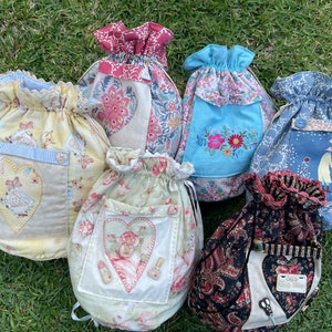 May include: Six drawstring bags made of fabric with various floral patterns and designs. The bags have a heart-shaped pocket on the front and are decorated with embroidery and appliques.