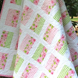 May include: A white quilt with a pink polka dot border. The quilt features a pattern of alternating pink floral and green gingham blocks, set against a white background. The quilt is draped over a green surface.