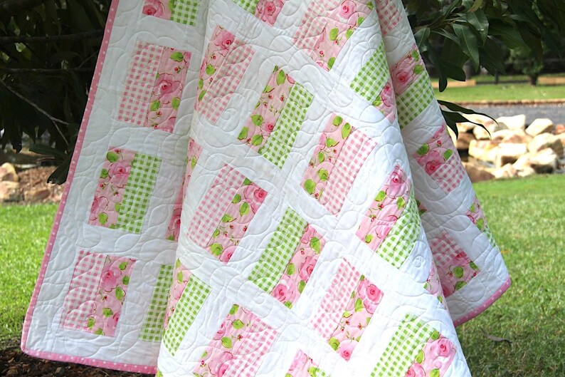May include: A white quilt with a pink polka dot border. The quilt features a pattern of alternating pink floral and green gingham blocks, set against a white background. The quilt is draped over a green surface.