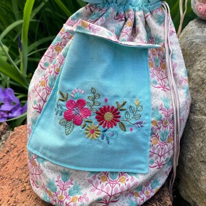 May include: A light blue drawstring bag with a floral pattern and a smaller light blue pocket with embroidered flowers in pink, red, and yellow.