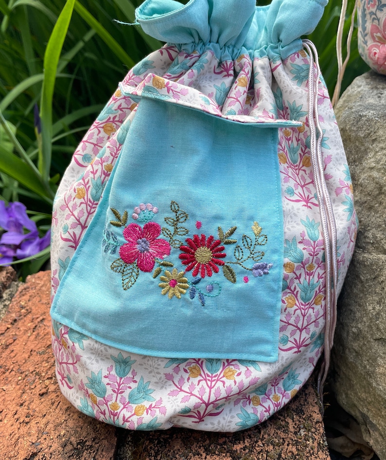 May include: A light blue drawstring bag with a floral pattern and a smaller light blue pocket with embroidered flowers in pink, red, and yellow.