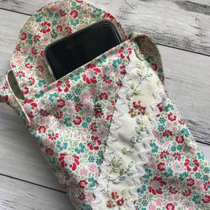 May include: A fabric phone pouch featuring a floral pattern in shades of red, pink, and teal on a cream background. The pouch has a flap closure with a silver snap button and a vertical cream-colored panel decorated with white rickrack trim.