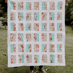 May include: A handmade quilt with a white background and rectangular blocks of various patterns. The blocks alternate between floral prints, solid tan, and solid teal. The quilt is held up against a green background.