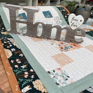 May include: A white wooden sign spelling "Home" with a white heart-shaped sign that says "Love the life you live" on a table runner with a floral pattern.