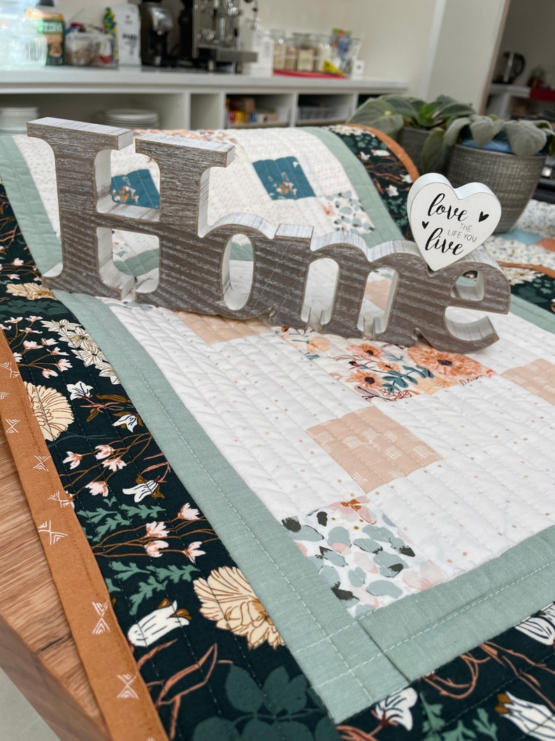 May include: A white wooden sign spelling "Home" with a white heart-shaped sign that says "Love the life you live" on a table runner with a floral pattern.
