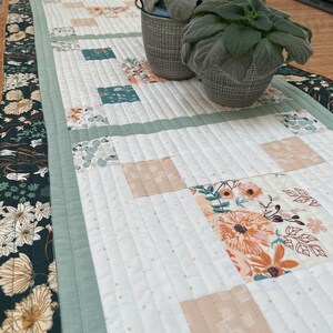 May include: A white quilted table runner with a green border and a floral pattern in the center. Two small potted plants are on the table runner.