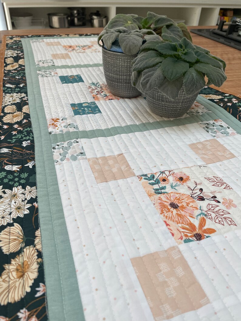 May include: A white quilted table runner with a green border and a floral pattern in the center. Two small potted plants are on the table runner.