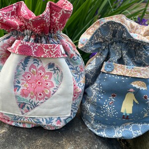May include: Two fabric drawstring bags with floral patterns. One bag is pink and blue with a heart-shaped patch. The other bag is blue with a small illustration of a person with an umbrella.