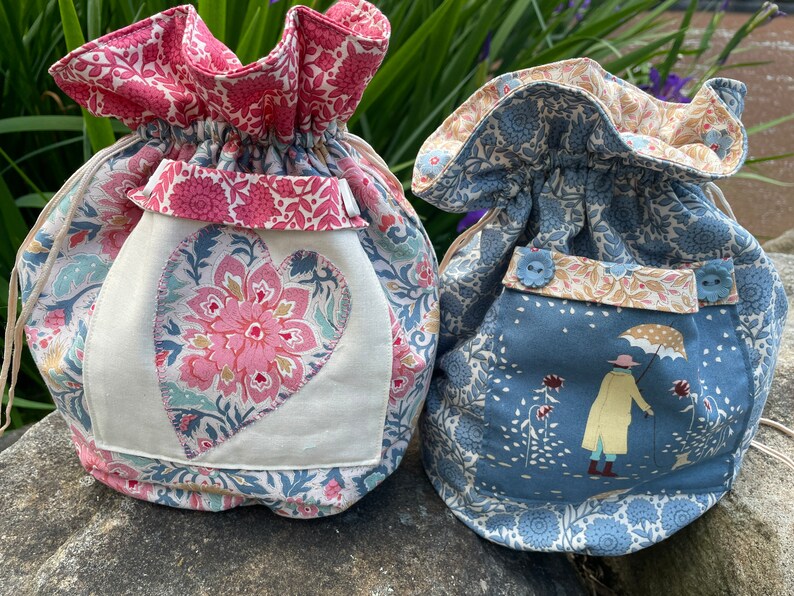 May include: Two fabric drawstring bags with floral patterns. One bag is pink and blue with a heart-shaped patch. The other bag is blue with a small illustration of a person with an umbrella.